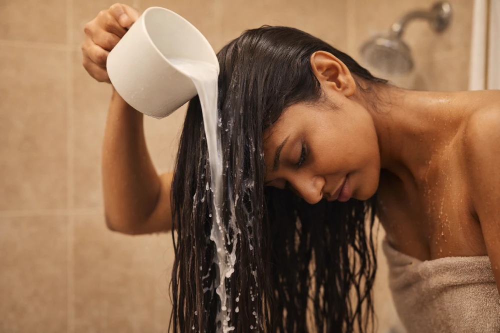 Woman pouring rice water rinse over dark hair in shower demonstrating correct application technique